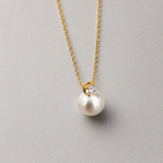 Lustre Necklace 316-kirurginteräksestä, 18K PVD-kultapinnoitteella, kullanvärinen kaulakoru helmellä ja kirkkaalla kivellä vaaleaa neutraalia taustaa vasten.