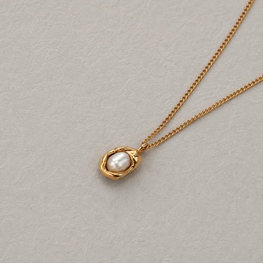 Oyster Necklace 316-kirurginteräksestä, 18K PVD-kultapinnoitteella, kullanvärinen kaulakoru pyöristetyllä helmiyksityiskohdalla vaaleaa neutraalia taustaa vasten.