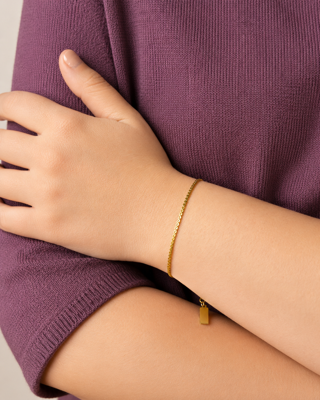 Ripple Bracelet i 316 kirurgiskt stål och 18k PVD-guld, ett nätt guldfärgat armband buret på handleden mot lila stickad textil.