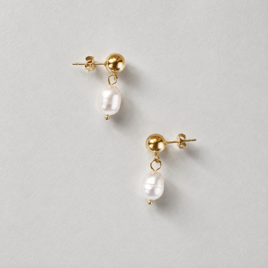 Satin Pearl Drop 316-kirurginteräksestä, 18K PVD-kultapinnoitteella, kullanväriset korvakorut riippuvilla helmillä vaaleaa neutraalia taustaa vasten.