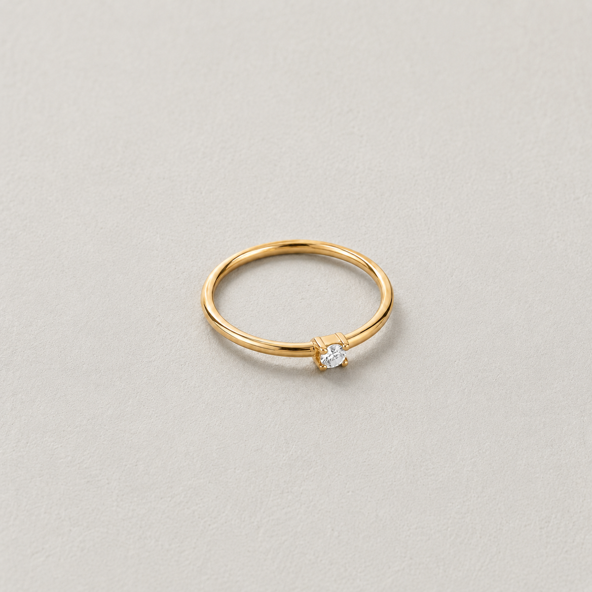 Dew Ring, kapea kullanvärinen sormus kirkkaalla kivellä vaalealla kivitaustalla.