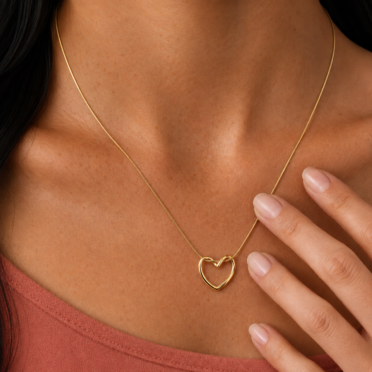 Echo Necklace 316-kirurginteräksestä, 18K PVD-kultapinnoitteella, kullanvärinen kaulakoru avoimella sydänriipuksella lämpimän terrakotanväristä toppia vasten.