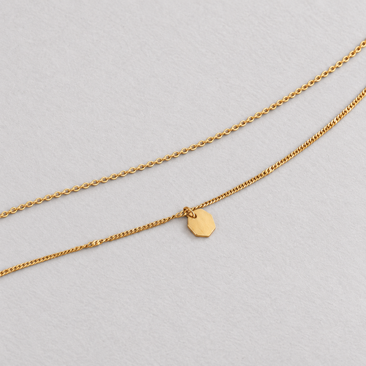 Facet Layer Necklace i guld med två tunna kedjor och facetterad berlock på ljus stenyta.
