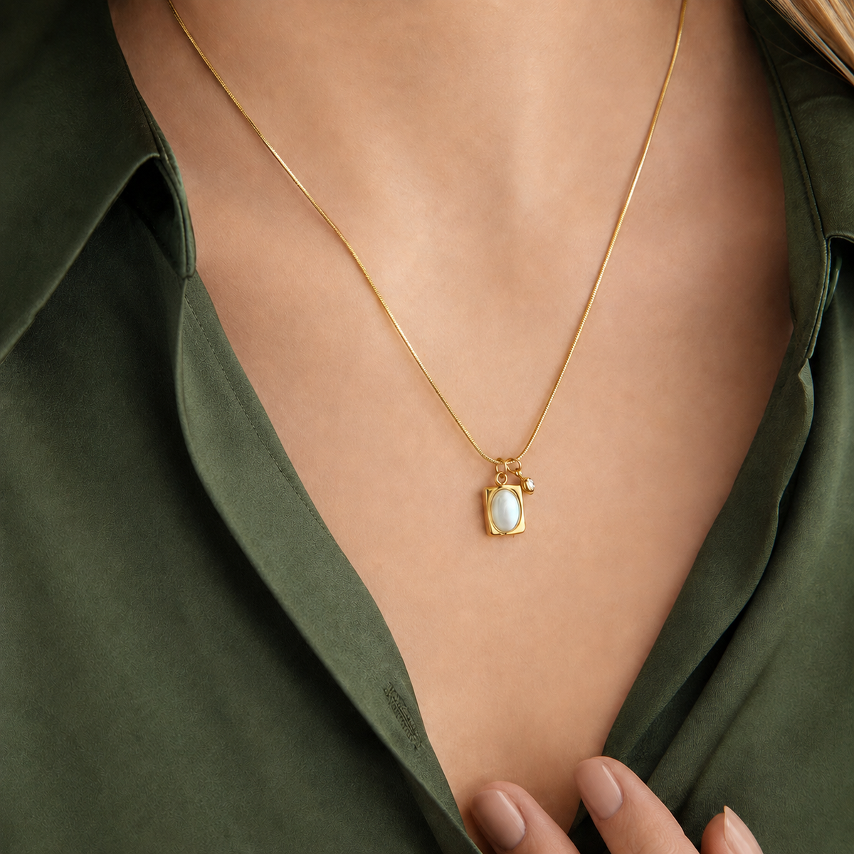 Halo Necklace i 316 kirurgiskt stål och 18k PVD-guld, ett guldfärgat halsband med ljust ovalt hänge buret på halsen mot mörkgrön skjorta.