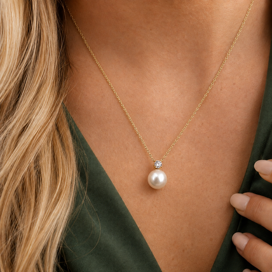 Lustre Necklace 316-kirurginteräksestä, 18K PVD-kultapinnoitteella, kullanvärinen kaulakoru helmellä ja kirkkaalla kivellä vihreää kangasta vasten.