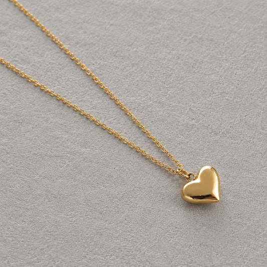 Pulse Necklace i 316 kirurgiskt stål och 18k PVD-guld, guldfärgat halsband med hjärtformat hänge mot ljus neutral bakgrund.