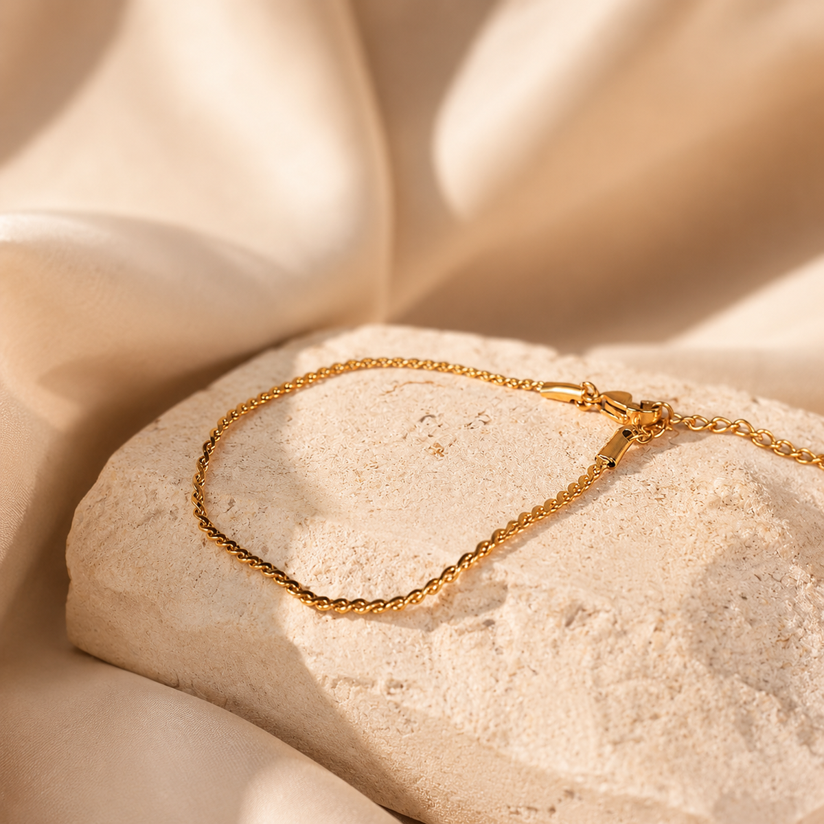Ripple Bracelet i 316 kirurgiskt stål och 18k PVD-guld, ett tunt guldfärgat armband på varm stenbakgrund med mjuk satäng.
