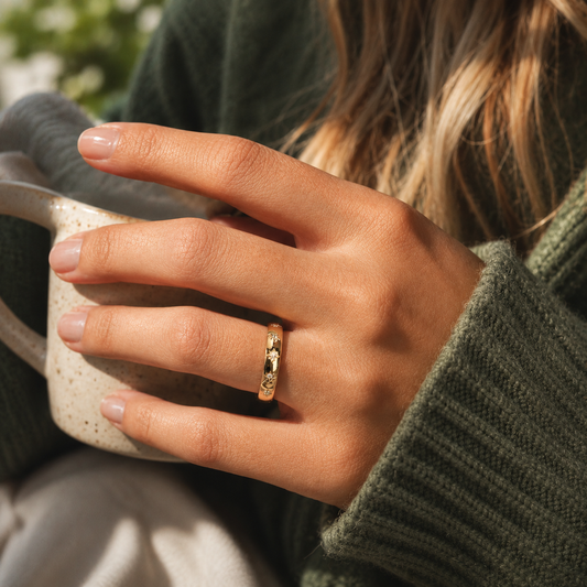 Sol Ring på hand, guldfärgad ring med små klara stenar och stjärnformade detaljer, fotograferad i varm miljö medan personen håller en keramikmugg.