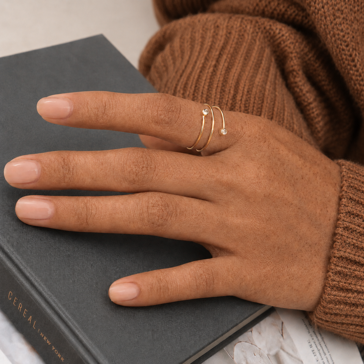 Spire Ring, en guldfärgad öppen ring med två små klara stenar, buren på handen ovanpå en bok tillsammans med en brun stickad tröja.