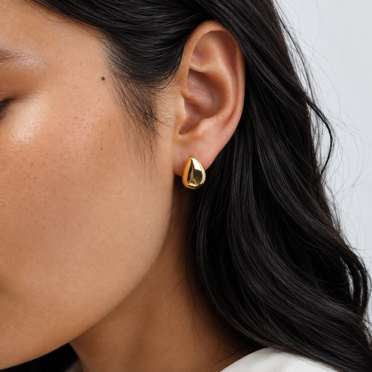 Stone Studs 316-kirurginteräksestä, 18K PVD-kultapinnoitteella, veistokselliset pisarakorvakorut korvissa vaaleaa neutraalia taustaa vasten.